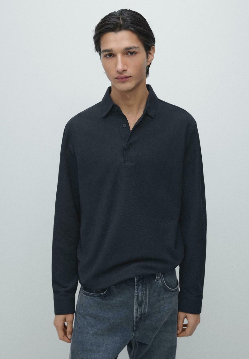 Marineblaues Langarm-Poloshirt mit Kragen, Knopfleiste und strukturierter Oberfläche, kombiniert mit dunklen Jeans. Modell wird von der Taille aufwärts gezeigt.