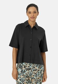 Camicia nera a maniche corte con collo e chiusura frontale a bottoni, realizzata in un tessuto liscio. Abbinata a pantaloni fantasia con disegni di foglie.
