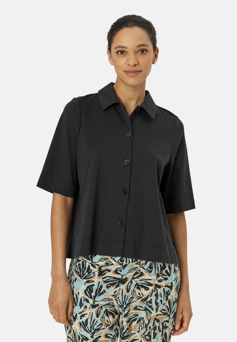 Camicia nera a maniche corte con collo e chiusura frontale a bottoni, realizzata in un tessuto liscio. Abbinata a pantaloni fantasia con disegni di foglie.