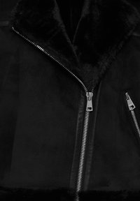 Chaqueta negra con cuello de piel sintética, textura de cuero suave y cremalleras metálicas a ambos lados, con un corte ligeramente oversize.