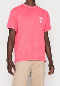 Man som har på sig en tvättad rosa t-shirt med kort ärm, med en liten vit ormlogo och texten "VIPER PIT" på vänster bröst, beige byxor.