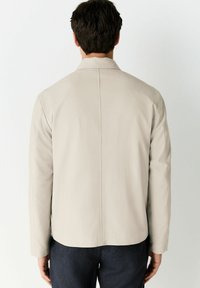 Chaqueta beige a medida con corte recto, mangas largas y una tela texturizada. Presenta una costura en la parte posterior y un diseño minimalista.