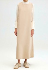Beige-mekko ilman hihoja, jossa on suora leikkaus, korkea pääntie ja sileä pinta. Pukeutunut kermaiseen pitkähiharaiseen paitaan, jossa on nappikoristeet.