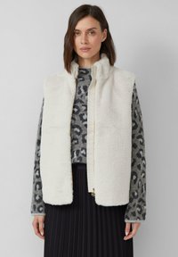 Eine Frau trägt einen weißen Kunstpelz-Westen über einem grauen Leopardenmuster-Pullover und einem schwarzen plissierten Rock, während sie vor einem schlichten grauen Hintergrund steht.