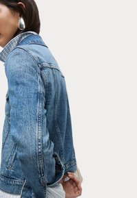 Modrá denimová bunda s klasickým střihem, s špicatým límcem, bočními kapsami a zapínáním na knoflíky. Textura je mírně vybledlá.