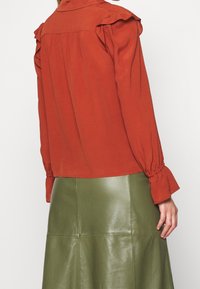 Rostfarbige Bluse mit Rüschenakzenten an den Schultern, kombiniert mit einem grünen Lederrock mit enganliegender Taille und ausgestellt geschnittenem Saum.