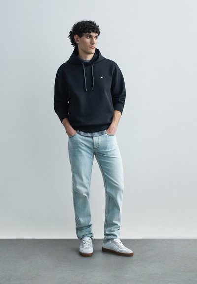 Melns kapuce ar aukliņām, gaiši zilas džinsa bikses un balti sporta apavi ar brūnām zolēm. Modelis stāv pret neitrālu fonu.