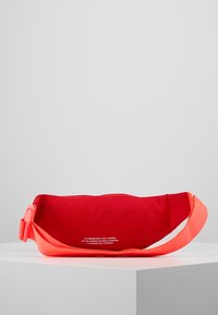 Červená látková taštička s jasně oranžovým nastavitelným popruhem. Obsahuje zipový otvor a tištěný text na přední straně. Plochý oválný tvar.