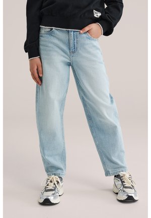 Személy világoskék, laza szabású farmernadrágot, fekete pulóvert szöveges folttal, valamint fehér, szürke és fekete vastag talpú sportcipőt visel, egyik kezét a zsebében tartja.