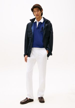 Tânăr purtând o jachetă bleumarin peste un polo albastru, pantaloni albi și mocasini maro închis, stând cu o mână în buzunar.