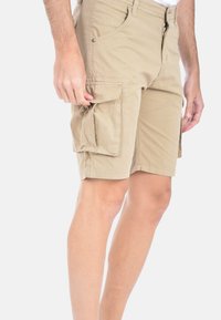 Pantaloni cargo beige realizzati in tessuto misto cotone, dotati di molteplici tasche, zip frontale e vestibilità comoda. Texture liscia con dettagli cuciti.