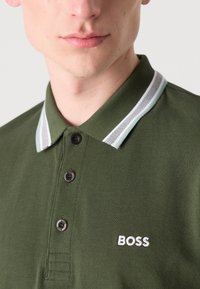 Mørkegrønn polo-skjorte med strukturert stoff, med krage i hvite og lys blå striper og svarte knapper. "BOSS"-logo i hvitt.