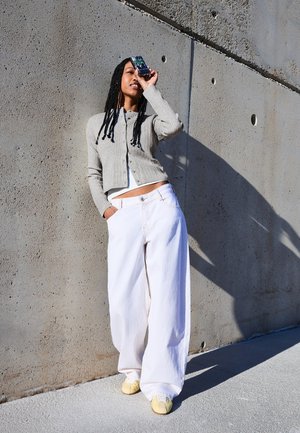 Frau mit langen Zöpfen steht vor einer Betonwand, hält ein Smartphone vor ihr Auge, trägt einen grauen Pullover und weite weiße Hosen.