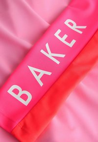 Žiarivo ružová a červená látka s bielym nápisom „BAKER“ vytlačeným veľkými písmenami v strede.