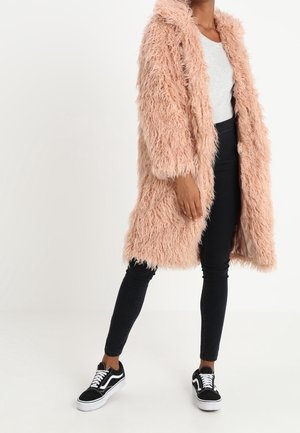 Person trägt einen langen, flauschigen rosa Mantel, weißes Oberteil, schwarze Skinny-Jeans und schwarz-weiße Turnschuhe und steht vor einem schlichten weißen Hintergrund.