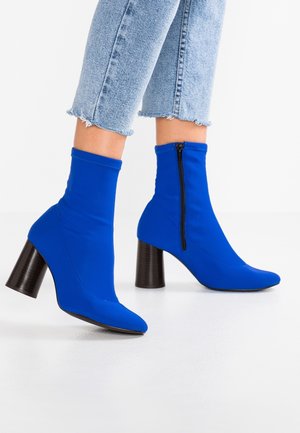 Personne portant des bottines à talons bleues vives avec des fermetures éclair latérales et un jean bleu clair raccourci avec des bords effilochés, debout sur un sol blanc.