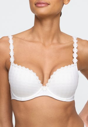 Vrouw draagt een witte beha met bloemenkantafwerking op de cups en bandjes, waarbij de bovenkant van de borst en de nek zichtbaar zijn tegen een effen achtergrond.