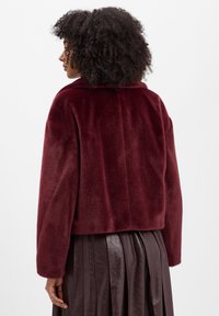 Vrouw met krullend donker haar, gekleed in een korte, fluwelen bordeauxrode jas en een geplooide donkerbruine leren rok, met de rug naar de camera gericht.