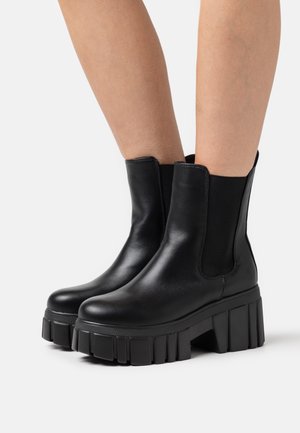 Bottines en cuir noir avec une plateforme épaisse et une semelle striée, dotées de panneaux latéraux élastiques pour un enfilage facile. Texture lisse, design épuré.
