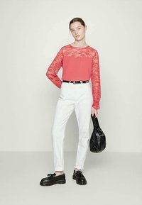 Top de renda coral com mangas longas, acompanhado por calças brancas de corte elegante. A modelo segura uma bolsa preta texturizada e veste sapatos pretos.