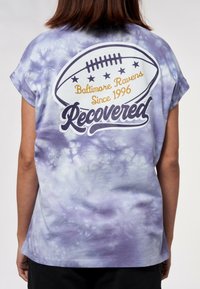 Lila tie-dye T-shirt med rullade korta ärmar. Har en fotbollsgraphic med "Baltimore Ravens Sedan 1996" och texten "Återvunnen".