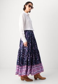 Jupe maxi évasée en marine avec un motif paisley, ornée d'un ourlet rose et blanc. Associée à une chemise blanche à boutons et des bottes chevilles marron.
