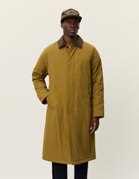 Olivno zeleni dolg plašč z gladko teksturo in rjavim žametnim ovratnikom, ki ima sprednji zadrgo in stranske žepe. Rjava kapa dodaja kontrast.