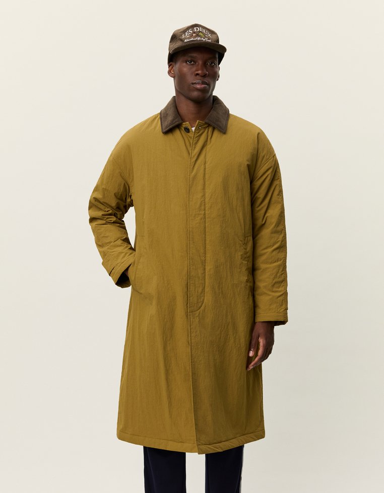 Olivno zeleni dolg plašč z gladko teksturo in rjavim žametnim ovratnikom, ki ima sprednji zadrgo in stranske žepe. Rjava kapa dodaja kontrast.