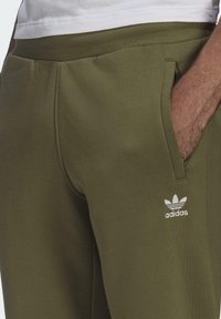 Mjukisbyxor i olivgrönt gjort av mjukt tyg, med sidofickor och en vit broderad Adidas-logotyp nära midjan.