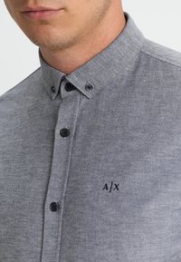 Szara koszula na guziki wykonana z fakturowanej tkaniny, z kontrastowymi czarnymi guzikami i małym wyszywanym logo "A/X" na piersi.