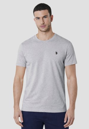 Uomo con capelli corti e scuri e occhi azzurri che indossa una T-shirt grigio chiaro a maniche corte con un piccolo logo nero sul petto.