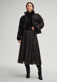 Mulher vestindo uma jaqueta acolchoada preta, suéter canelado preto, saia longa estampada e botas pretas até ao joelho contra um fundo simples.
