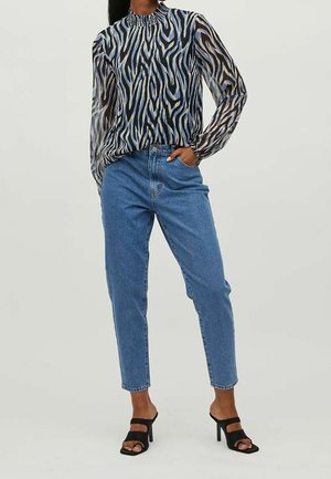 Kobieta ubrana w przezroczystą bluzkę w zebra nadruk, z długim rękawem, wysokim stanem niebieskich dżinsów i czarne sandały na wysokim obcasie z odkrytymi palcami, stoi z dłonią w kieszeni.