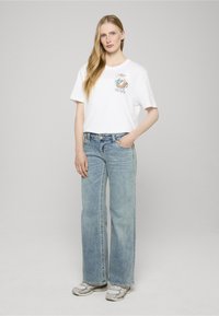 Bílé bavlněné tričko s grafickým motivem na hrudi, v kombinaci se světle modrými širokými džínami a šedobílými teniskami.