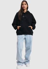 Sort hættetrøje med forlommes, afslappet pasform og hvidt logo; parret med lyseblå brede jeans og hvide sneakers.