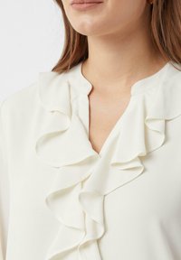 Biała bluzka z dekoltem w kształcie V, z wyraźnymi falbanami biegnącymi wzdłuż przodu oraz miękką, gładką teksturą materiału.