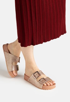Feet wearing open-toe glittery rose gold sandals with two buckle straps, shown with a red pleated skirt.