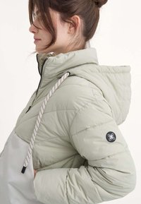 Perfil de una mujer que lleva una chaqueta acolchada verde clara con capucha, que muestra una vista lateral y los detalles de la chaqueta.