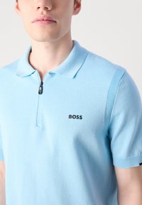 Camisa polo azul clara feita de tecido tricotado, com fecho de zipper e mangas curtas. O logótipo "BOSS" é visível no lado esquerdo do peito.
