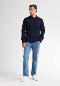 Marineblauw overhemd met knopen en een ruitpatroon, gecombineerd met lichtblauwe jeans en witte sneakers. Het overhemd heeft een zak en een kraag.