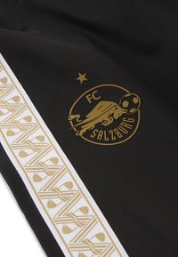 Sorte sportsbukser med et guld FC Salzburg-logo og en fodbold, hvid sidelinje med indviklede guldmønstrede detaljer.