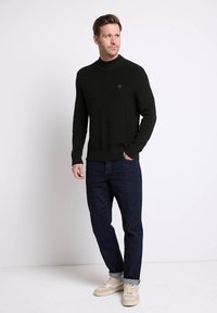 Pull à côtes noir avec un col montant, associé à un jean bleu foncé et des baskets de couleur claire. Présente un petit logo sur le pull.