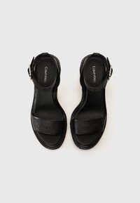 Black sandals with adjustable ankle strap, featuring wide front band embossed with "Calvin Klein." Smooth leather texture and flat sole.