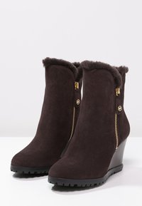 Bruna kilklackade ankelstövlar i mocka med dragkedja, fuskpälskant upptill och texturerade gummisulor för grepp. Guldiga metalldetaljer.