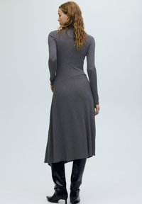 Hajlított hajú nő, szürke testhez simuló hosszú ujjú midi ruhát és fekete térdig érő, sarkú csizmát viselve, háttal áll egy egyszerű háttér előtt.