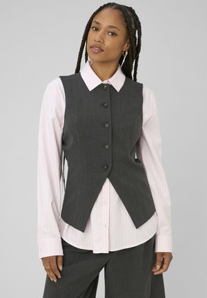 Femme aux cheveux tressés portant des boucles d’oreilles créoles en or, une chemise rose clair à rayures boutonnée, et un gilet sans manches et un pantalon gris foncé.