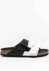 Black and white two-band sandal with adjustable buckles, cork footbed, and textured black rubber outsole. Minimalist design for casual wear.