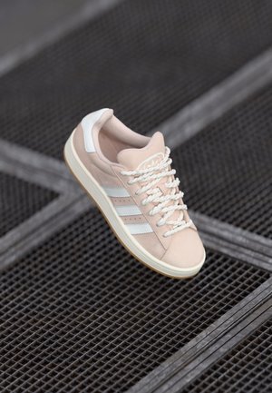 Jasnoróżowy sneaker Adidas z białymi paskami i sznurowadłami, przedstawiony na tle ciemnej metalowej kraty.