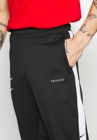 Svarta sportbyxor med vita sidostripes och "SWOOSH"-logotyp. Har resårmidja och fickor. Tillverkad av ett glatt, lätt material.