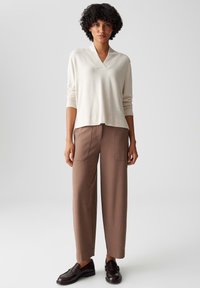Lichtbeige, lange mouwen V-hals trui gecombineerd met losse, bruine broek met zijzakken. De schoenen zijn donkerbruin, gesloten teen met versiering.